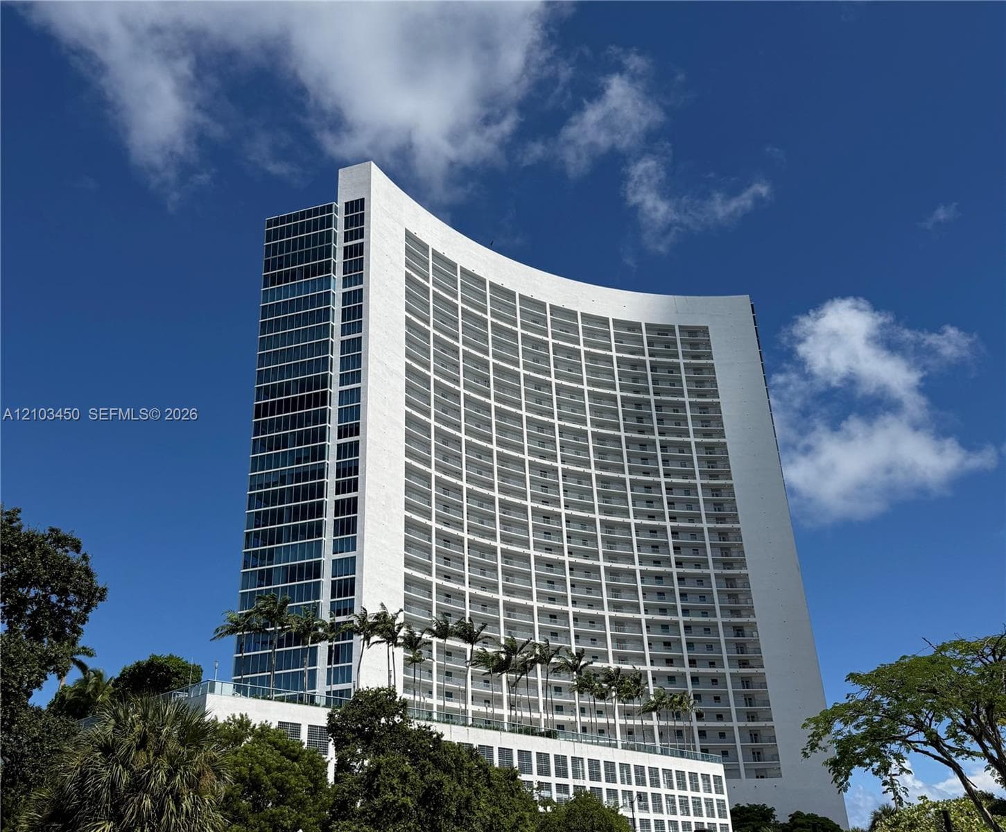 Curved high-rise exterior featuring white architectural concrete facade, extensive glass curtain wall, and stacked private balconies