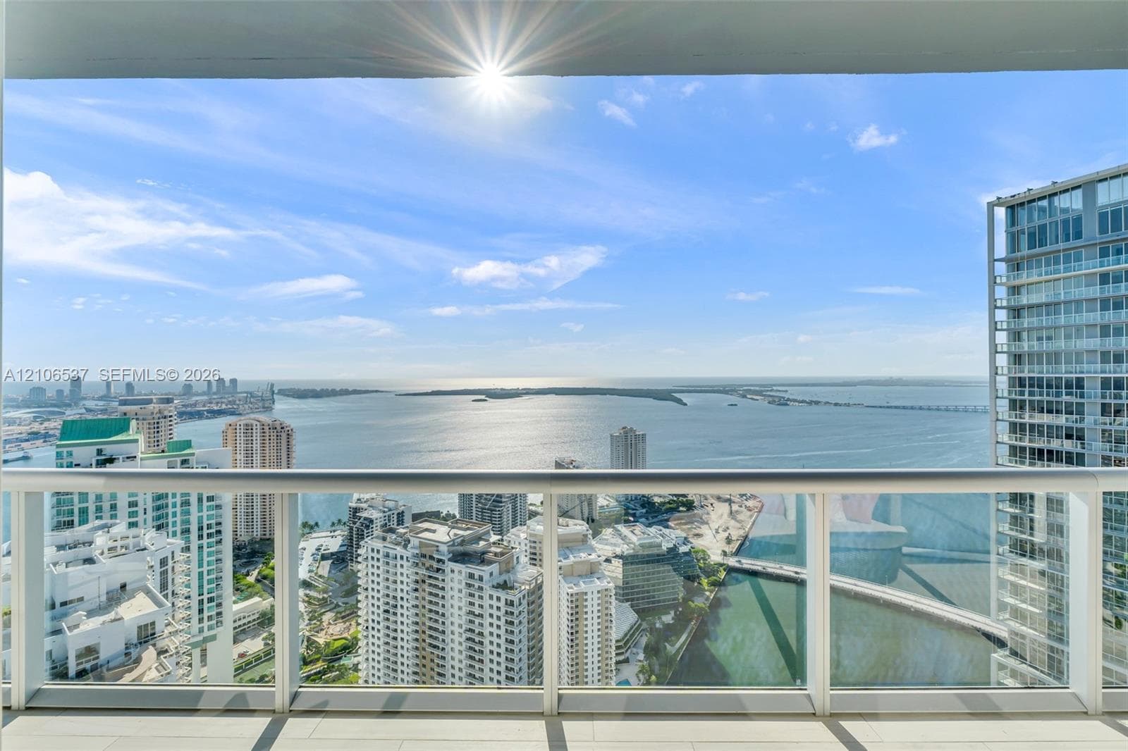 Covered balcony with a glass-panel railing, tile flooring, and expansive water and urban skyline backdrop