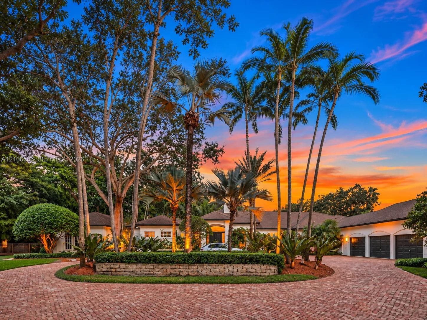 Circular brick paver driveway, mature landscaping with palm trees, and multi-car garage