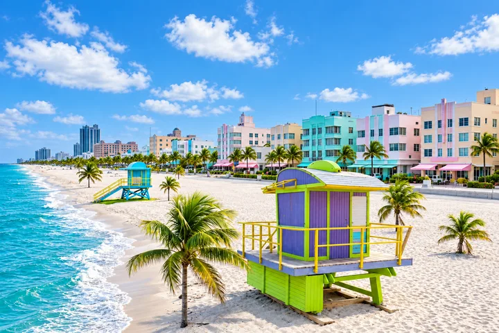 Coastal scene with colorful lifeguard cabins, palm trees, pastel Art Deco buildings and turquoise surf under a blue sky